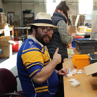 Damien, a trainee, is wearing a blue and yellow shirt and a straw hat. he is sat at his desk working on a task and is happy as he is showing his thumb up.